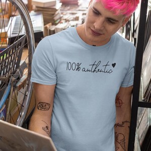 May include: A light blue t-shirt with the text "100% authentic" and a small black heart. The person wearing the shirt has tattoos on their arms.