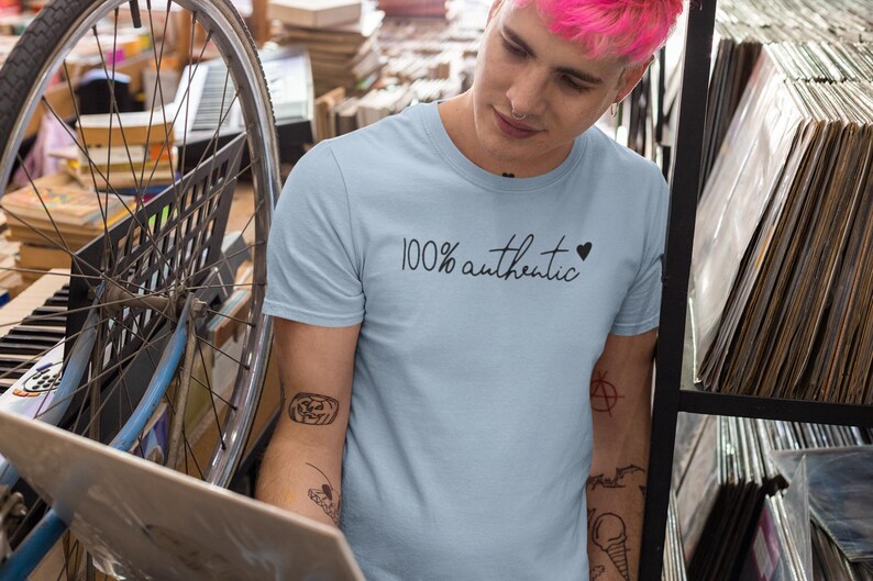 May include: A light blue t-shirt with the text "100% authentic" and a small black heart. The person wearing the shirt has tattoos on their arms.