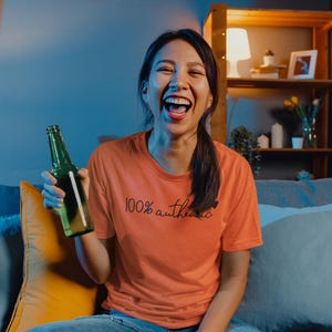 May include: A woman wearing a peach colored t-shirt with the text "100% authentic" printed on it. She is holding a green glass bottle and laughing. She is sitting on a couch with blue and yellow pillows.