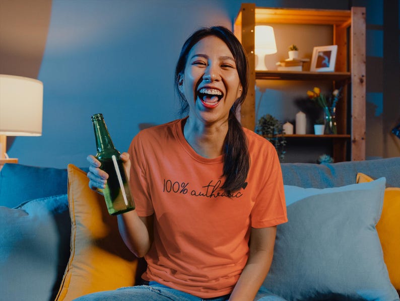 May include: A woman wearing a peach colored t-shirt with the text "100% authentic" printed on it. She is holding a green glass bottle and laughing. She is sitting on a couch with blue and yellow pillows.
