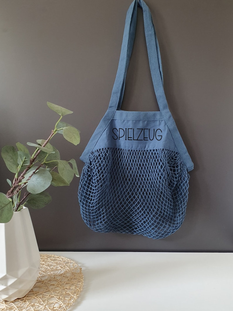May include: A blue canvas tote bag with a mesh bottom. The bag has a black embroidered word "SPIELZEUG" on the front.