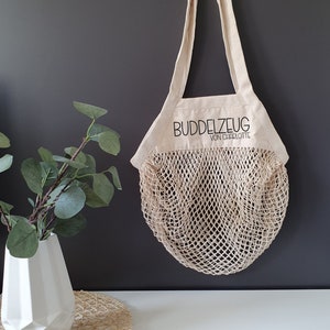 May include: A beige canvas tote bag with a mesh net bottom. The bag has a black embroidered logo that says "Buddelzeug von Charlotte".