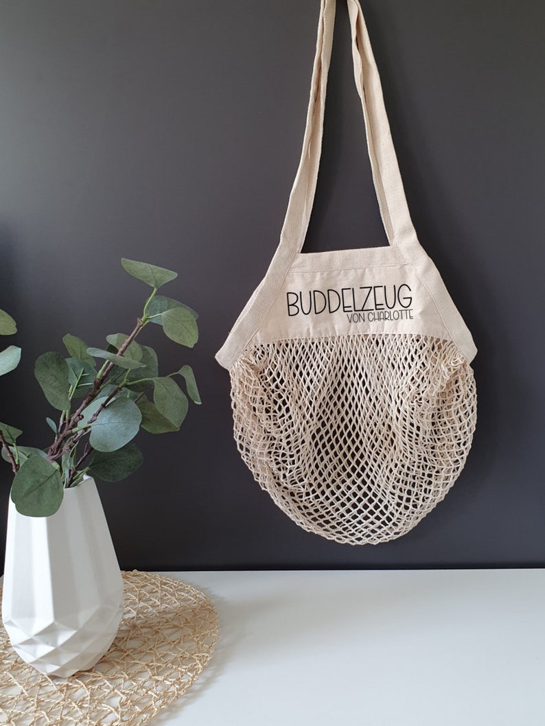 May include: A beige canvas tote bag with a mesh net bottom. The bag has a black embroidered logo that says "Buddelzeug von Charlotte".
