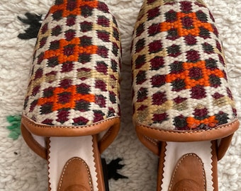 May include: A pair of brown leather slippers with a pointed toe and a colorful geometric pattern. The slippers have a white leather lining and a brown leather sole. The brand name "IA" is embossed on the sole.