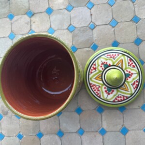 May include: A green ceramic jar with a lid. The lid features a colourful floral design with red, yellow, and green accents. The jar is empty and the inside is visible.