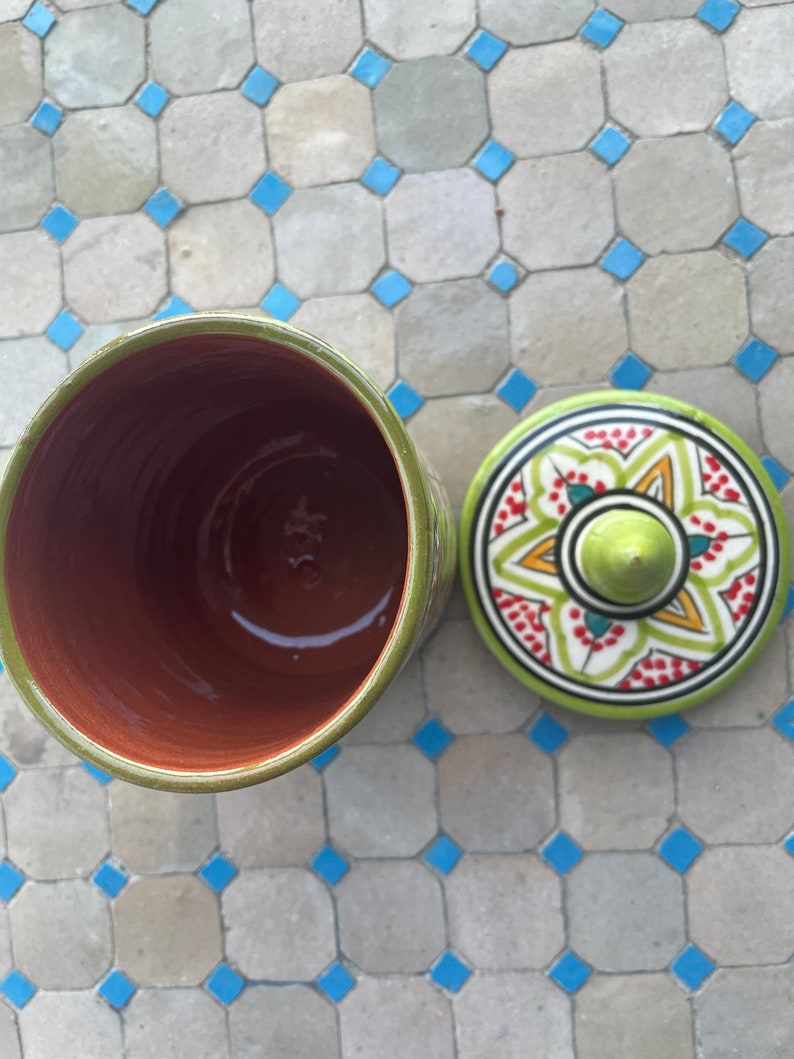 May include: A green ceramic jar with a lid. The lid features a colourful floral design with red, yellow, and green accents. The jar is empty and the inside is visible.