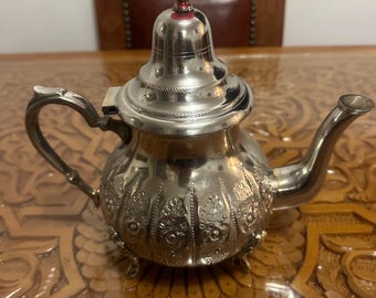 May include: A silver teapot with intricate floral designs and a red knob on the lid. The teapot has a rounded body and a spout on the side. It is sitting on a wooden table with a carved pattern.