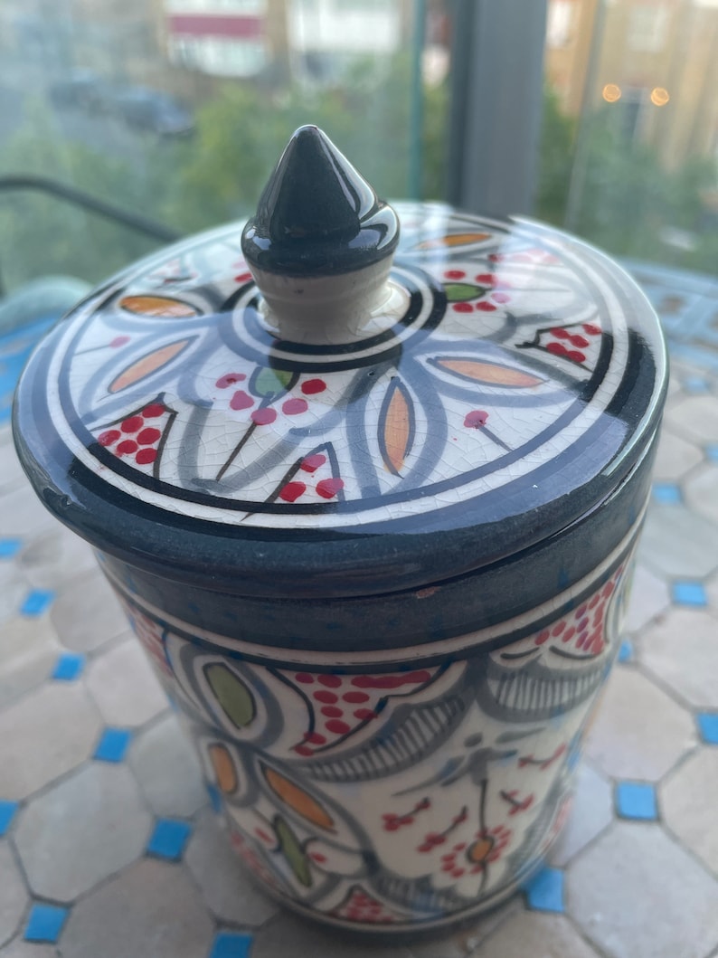 May include: A ceramic jar with a lid featuring a colourful floral design in shades of blue, green, red, orange, and white. The lid has a pointed top.