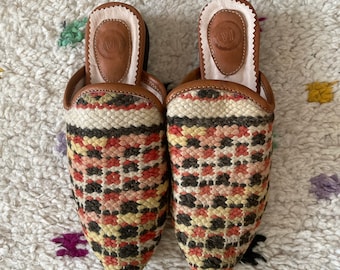 May include: A pair of pointed-toe, woven leather slippers with a geometric pattern in shades of red, pink, yellow, and black. The slippers have a brown leather heel and a white leather lining.
