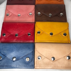 Puede incluir: Ocho carteras de cuero en diferentes colores, cada una con cierre de solapa y tres broches de presión plateados. Las carteras están dispuestas en dos filas de cuatro, con los colores alternando.