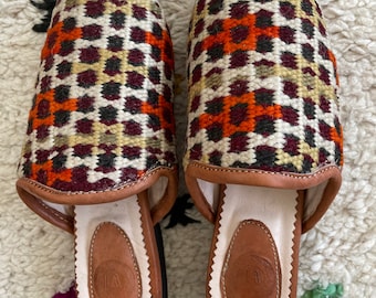 May include: A pair of patterned slippers with a geometric design in orange, burgundy, black, and cream. The slippers have a leather trim and a leather insole. The soles are black.