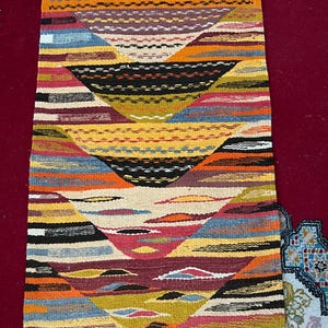 Op de afbeelding: Een rechthoekig, handgeweven tapijt met een geometrisch ontwerp. Het tapijt heeft een levendige mix van kleuren, waaronder oranje, geel, blauw en zwart, gerangschikt in een reeks chevron-achtige patronen. Het tapijt heeft een gefranjerde rand en is ongeveer 60 cm breed en 122 cm lang.
