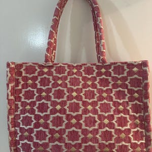 Könnte beinhalten: Eine rosa Tragetasche mit einem geometrischen Muster in Weiß und Gold. Die Tasche hat zwei Griffe und ist aus einem strukturierten Stoff gefertigt. Das Muster ist ein sich wiederholendes Design aus sternförmigen Formen.