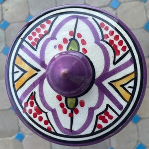 May include: A ceramic lid with a purple, white, yellow, and red geometric design. The lid has a purple knob on top.