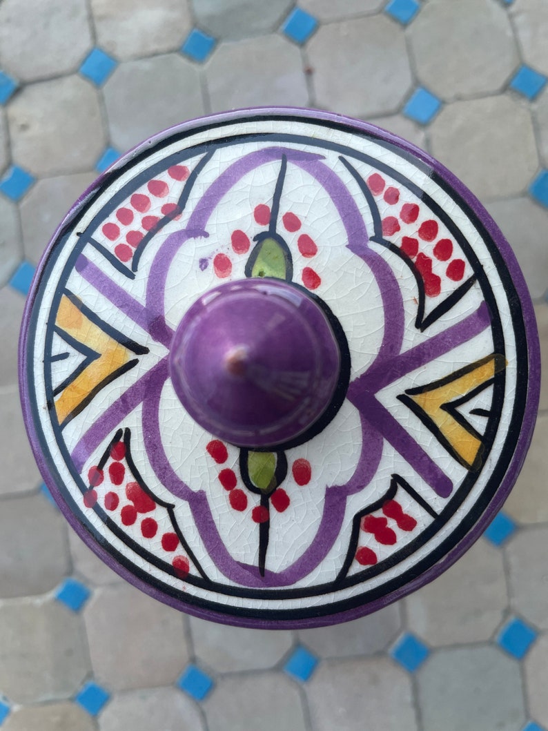 May include: A ceramic lid with a purple, white, yellow, and red geometric design. The lid has a purple knob on top.