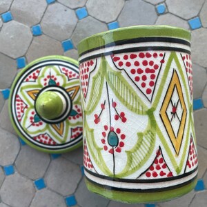 May include: A green ceramic jar with a lid, both decorated with intricate geometric patterns in red, yellow, and blue. The jar has a white background and a glossy finish.