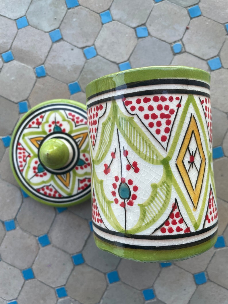 May include: A green ceramic jar with a lid, both decorated with intricate geometric patterns in red, yellow, and blue. The jar has a white background and a glossy finish.