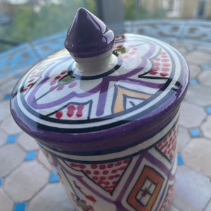 May include: A white ceramic jar with a purple lid and intricate geometric patterns in red, orange, and purple. The jar has a traditional Moroccan design.