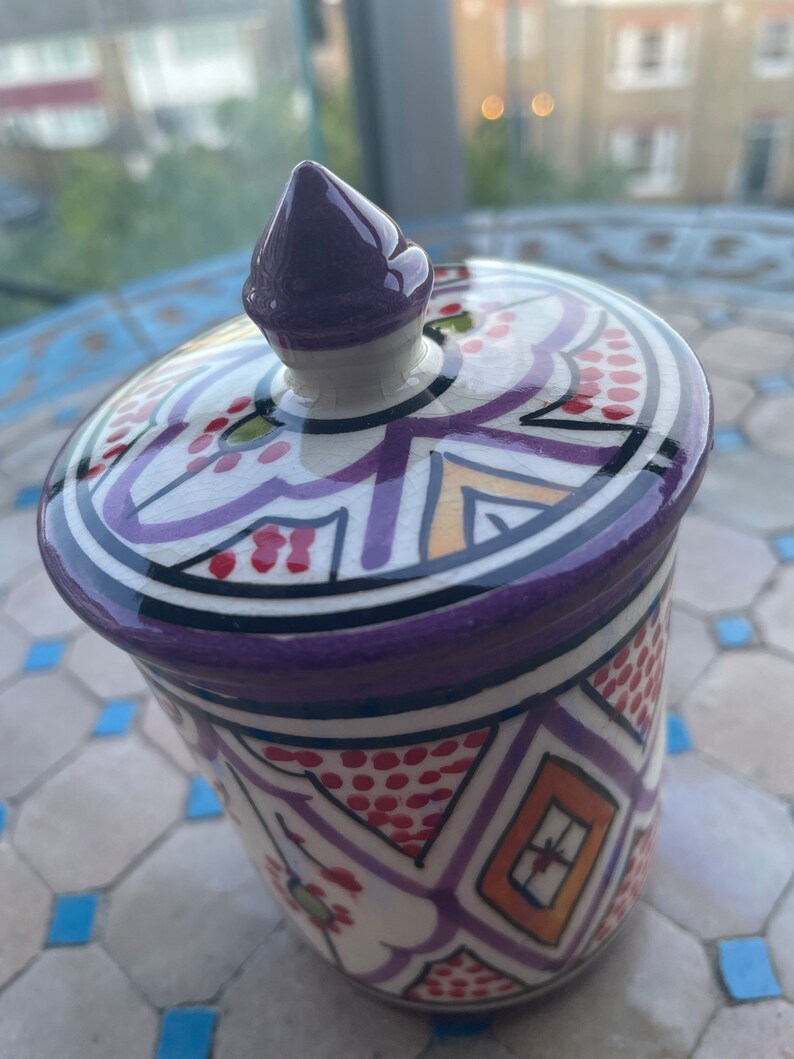 May include: A white ceramic jar with a purple lid and intricate geometric patterns in red, orange, and purple. The jar has a traditional Moroccan design.