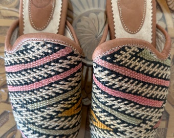 May include: A pair of black, pink, and beige woven slippers with pointed toes. The slippers have a brown leather lining and a white leather insole.
