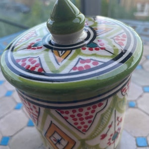 May include: A green ceramic jar with a lid. The jar has a white background with red, green, yellow, and black geometric patterns. The lid has a green knob.