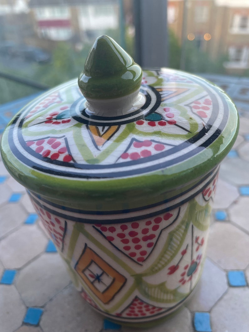 May include: A green ceramic jar with a lid. The jar has a white background with red, green, yellow, and black geometric patterns. The lid has a green knob.