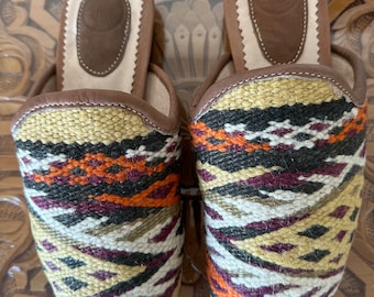 May include: A pair of colorful woven slippers with a geometric pattern in shades of orange, red, black, white, and brown. The slippers have a leather lining and a brown leather insole.