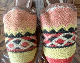 May include: A pair of pointed-toe slippers with a colorful woven pattern in shades of pink, red, yellow, black, and white. The slippers have a brown leather insole and a white trim around the top.