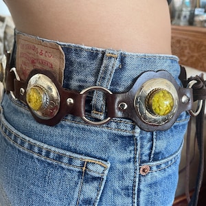 May include: A brown leather belt featuring circular silver-toned metal plates with embossed patterns and central yellow textured circular inlays. The belt is constructed with silver-toned metal rings connecting the leather segments and silver rivets.