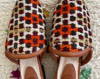 May include: A pair of brown leather and woven fabric slippers with a geometric pattern in shades of orange, red, black, and white. The slippers have a pointed toe and a flat sole.