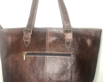 Puede incluir: Un bolso tote de cuero marrón con dos asas superiores y un bolsillo con cremallera en la parte delantera. El bolso tiene costuras blancas y herrajes dorados.