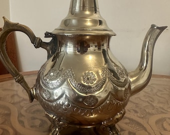 May include: A silver teapot with an ornate design and a red knob on the lid. The teapot has three feet and is sitting on a wooden table.