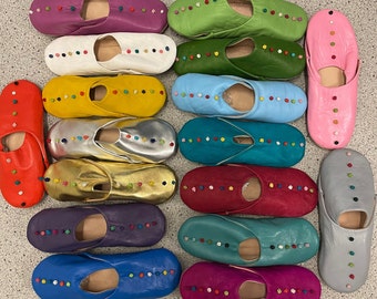 May include: A collection of 20 colorful leather slippers with embroidered details. The slippers are arranged in two rows, with each row containing 10 slippers. The slippers are all different colors, including red, blue, green, yellow, pink, and purple. The slippers are all decorated with small, colorful beads.
