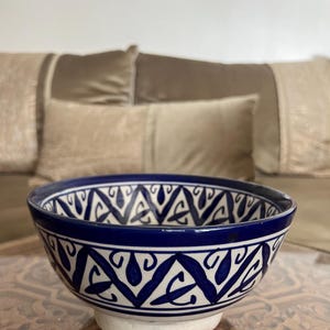 May include: A ceramic bowl with a white base and a deep blue rim. The bowl features a repeating white and blue geometric pattern around the exterior. The bowl is sitting on a wooden table.