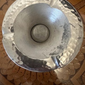 May include: A silver ashtray with a hammered finish and a round, indented center. The ashtray is sitting on a brown surface.