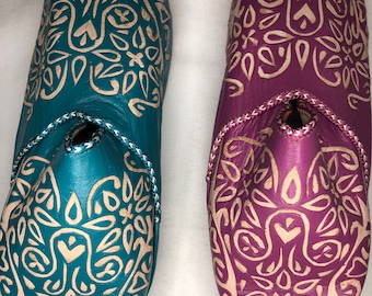 May include: Two pairs of pointed leather shoes with intricate white patterns on a teal and purple background.