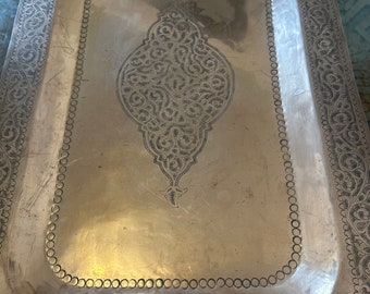 Puede incluir: Una bandeja rectangular de plata con un diseño ornamentado. La bandeja tiene un borde elevado con un patrón repetitivo de círculos. El centro de la bandeja presenta un gran diseño intrincado.