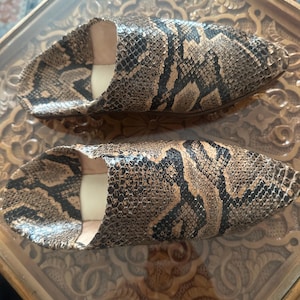May include: A pair of snake-print leather slippers. The slippers feature a pointed toe design and a neutral color palette of brown, black, and beige. The interior is a light beige color.
