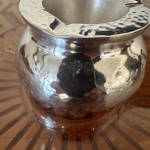 May include: A silver hammered metal ashtray with a round, wide base and a slightly flared top.