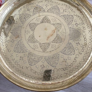 Può includere: Un vassoio rotondo in metallo dorato con un design in rilievo intricato. Il design presenta un'esplosione di stelle centrale circondata da motivi geometrici ripetitivi.