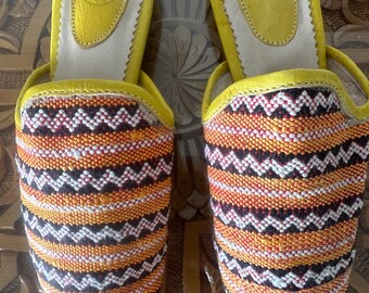 May include: A pair of yellow leather slippers with a pointed toe and a woven pattern in orange, black, and white. The slippers have a yellow leather insole with a white border and a small yellow leather tag with the letter "M" on it.