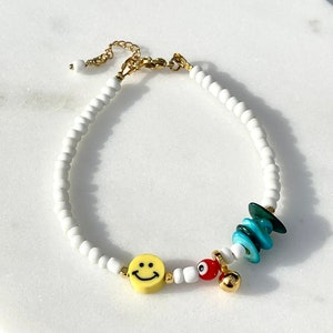 Puede incluir: Pulsera de cuentas blancas con cadena y cierre dorados. La pulsera presenta un dije de carita sonriente amarilla, detalles de piedras turquesas y verdes, un dije de ojo turco rojo y un dije de campana dorada.