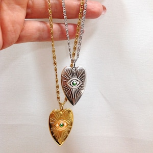 Peut inclure: Deux colliers pendentifs en forme de cœur. L'un est doré, l'autre argenté. Chaque pendentif présente un motif d'œil avec une gemme verte au centre. Les pendentifs sont suspendus à de fines chaînes.