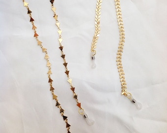 May include: Gold-colored eyeglass chains with different designs. One chain features triangle-shaped links, while the other has a chevron pattern. The chains have clear rubber loops for attaching to glasses and are displayed on a white background.