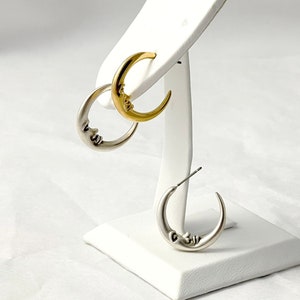 May include: A pair of crescent moon earrings, one gold-toned and one silver-toned, displayed on a white jewelry stand. Each earring features a smiling face design. The earrings are a celestial-themed accessory.