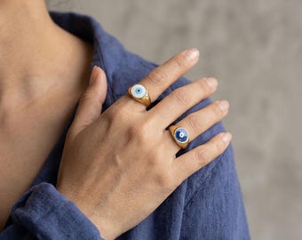 May include: Two gold-toned signet rings featuring circular evil eye motifs. One ring displays a white background with a blue and black center, while the other ring features a deep blue background with a white and black center eye design.
