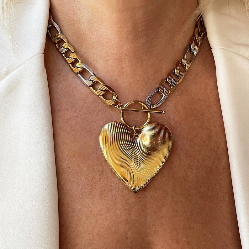 Op de afbeelding: Goudkleurige hartvormige hangerketting met een getextureerd oppervlak. De ketting heeft een dikke ketting met gouden en zilveren schakels. De hartvormige hanger is bevestigd aan een cirkelvormige sluiting en een staafsluiting. De ketting wordt gedragen over een witte blazer.