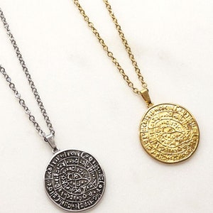 Peut inclure: Deux colliers avec des pendentifs ronds. L'un est argent&eacute;, l'autre dor&eacute;. Les deux pendentifs pr&eacute;sentent un motif grav&eacute; avec des symboles et du texte. Les colliers sont pr&eacute;sent&eacute;s sur une surface blanche.