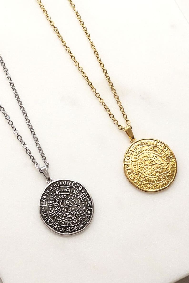 Peut inclure: Deux colliers avec des pendentifs ronds. L'un est argent&eacute;, l'autre dor&eacute;. Les deux pendentifs pr&eacute;sentent un motif grav&eacute; avec des symboles et du texte. Les colliers sont pr&eacute;sent&eacute;s sur une surface blanche.