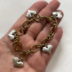 Könnte beinhalten: Ein goldfarbenes Gliederarmband mit silbernen Herzanhängern. Das Armband hat einen Knebelverschluss und eine Mischung aus ovalen und runden Gliedern. Die silbernen Herzen haben eine strukturierte Oberfläche.